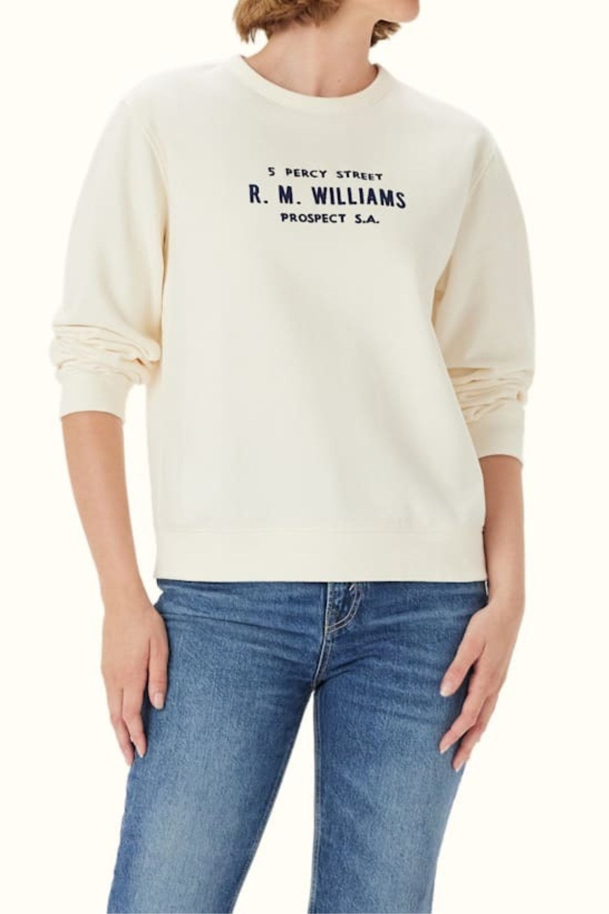 A person models the RM Williams Heritage Brushed Sweatshirt—cream, ribbed, crew neck with "5 Percy Street R.M. Williams Prospect S.A." printed in black—paired with blue jeans, head slightly turned and partially out of view.