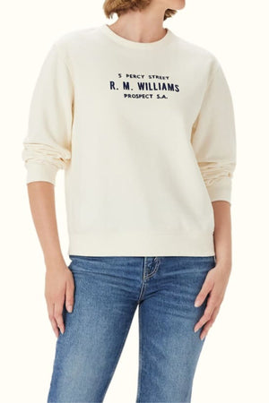 A person models the RM Williams Heritage Brushed Sweatshirt—cream, ribbed, crew neck with "5 Percy Street R.M. Williams Prospect S.A." printed in black—paired with blue jeans, head slightly turned and partially out of view.