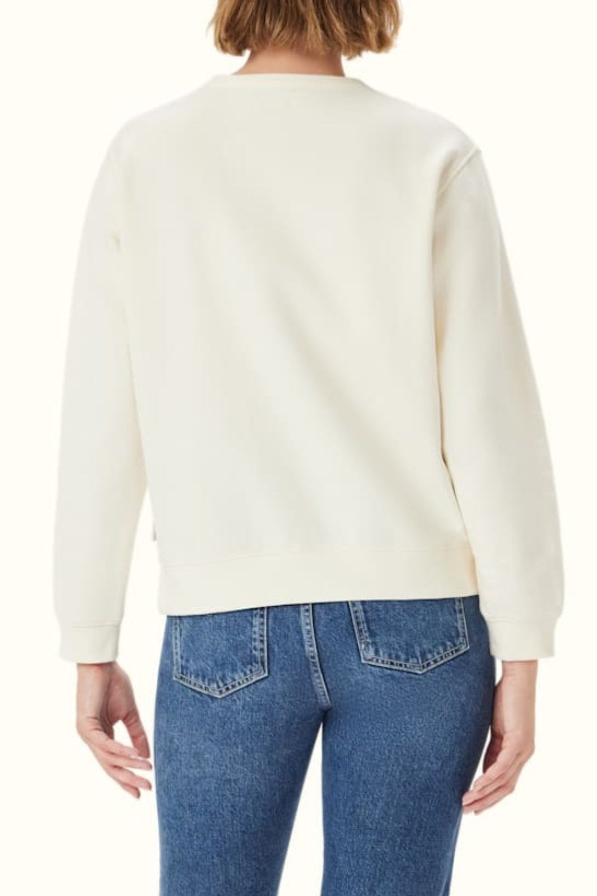 A person with short light brown hair is shown from the back, wearing the RM Williams Heritage Brushed Sweatshirt in cream with a ribbed crew neck and blue jeans, set against a neutral background.