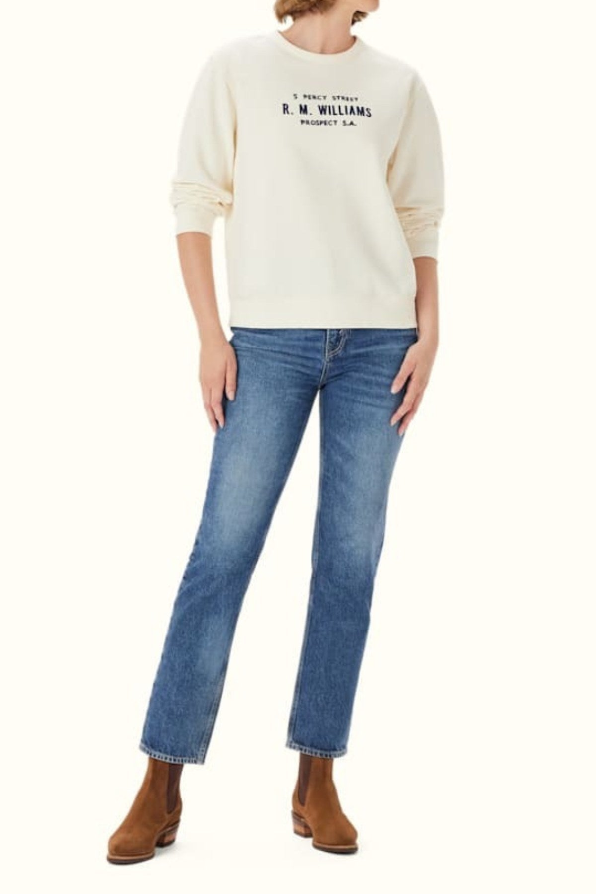 A person wearing an RM Williams Heritage Brushed Sweatshirt in cream with a ribbed crew neck, along with blue jeans and brown ankle boots, stands against a plain light background.