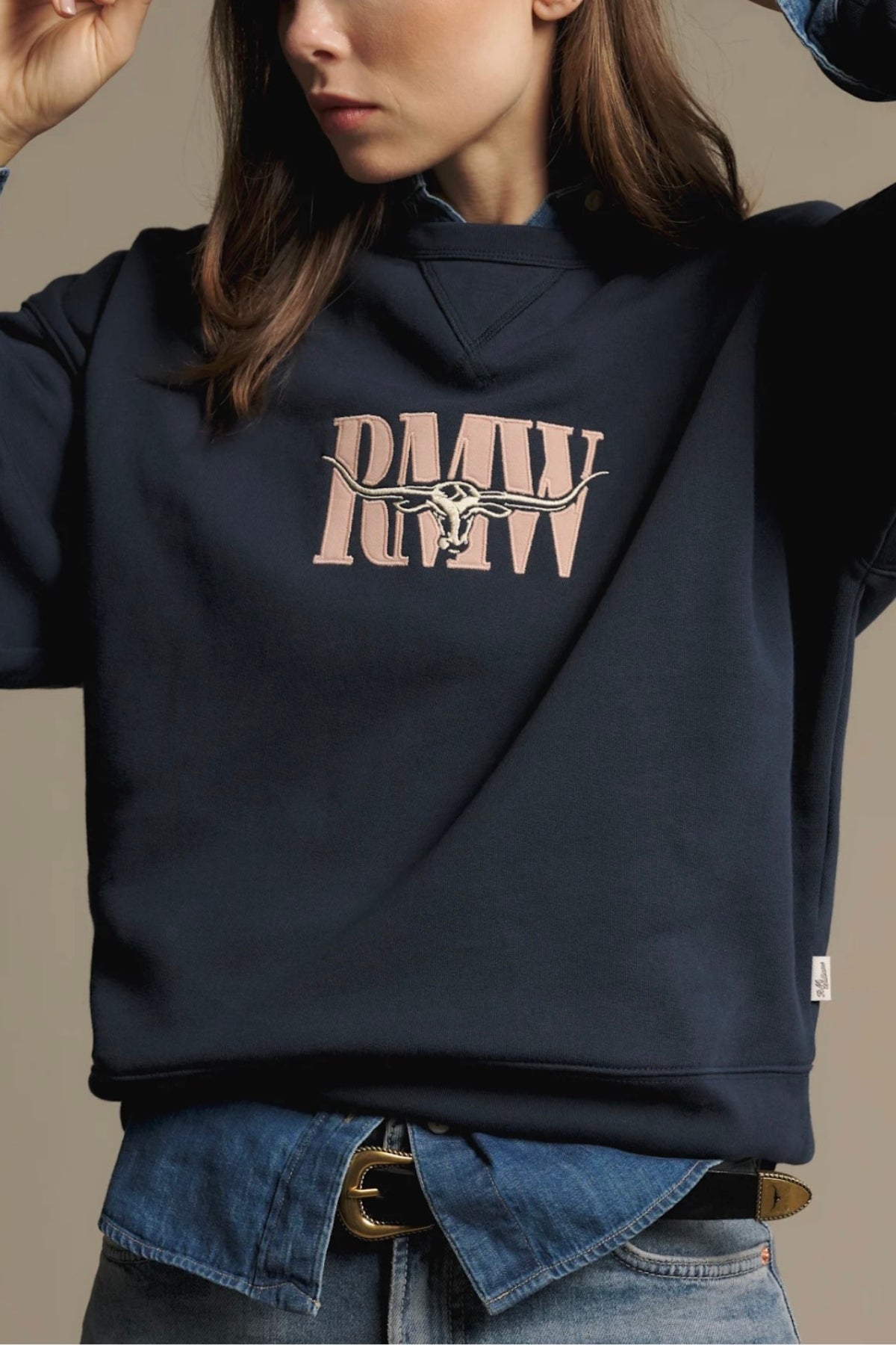 Wearing the RM Williams Heritage Easy Sweatshirt—a navy crew neck featuring "RMW" lettering and a horse-and-rider graphic—layered over a denim shirt, the woman poses with one hand near her face, highlighting the heritage branding while only the lower half of her face is visible.