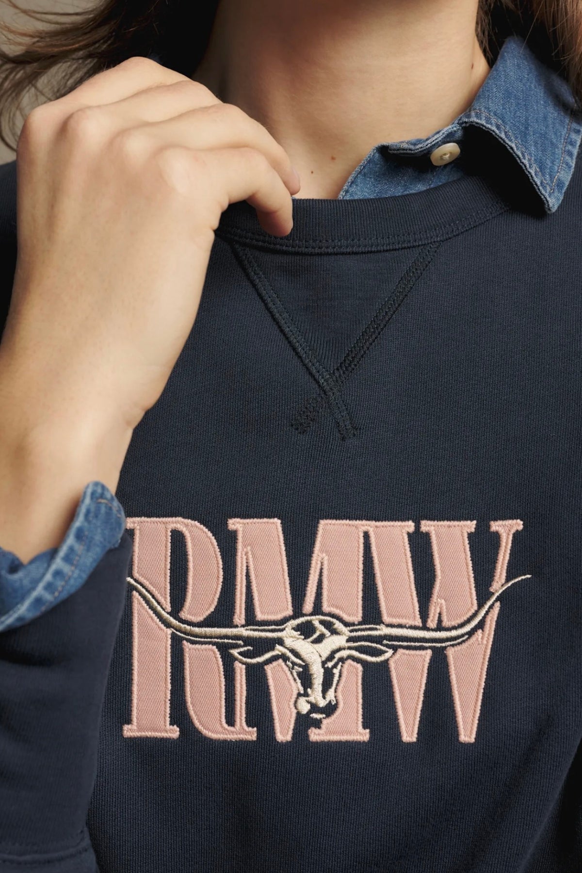 Wearing the RM Williams Heritage Easy Sweatshirt—a dark crew neck sweatshirt embroidered with "RMW" and a longhorn skull—layered over a blue denim collared shirt, the person showcases heritage branding with their hand resting near their chin.