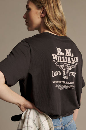 A woman stands with one hand on her hip and holding a plaid shirt, wearing the RM Williams Kandos Tee—a relaxed fit t-shirt crafted from premium Australian cotton, featuring R.M. Williams branding and a bold longhorn graphic with text on the back.