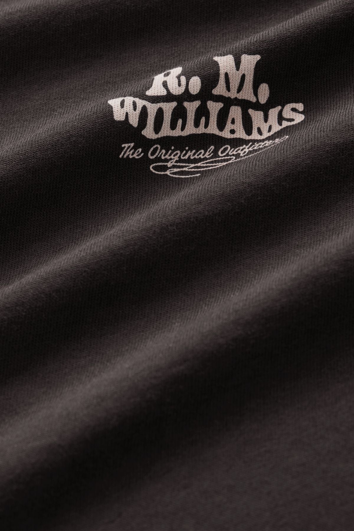 Close-up of the RM Williams Kandos Tee in black, showcasing a branded print with white text reading “R.M. WILLIAMS The Original Outfitters” in a stylized font on slightly wrinkled fabric for added texture.