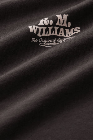 Close-up of the RM Williams Kandos Tee in black, showcasing a branded print with white text reading “R.M. WILLIAMS The Original Outfitters” in a stylized font on slightly wrinkled fabric for added texture.