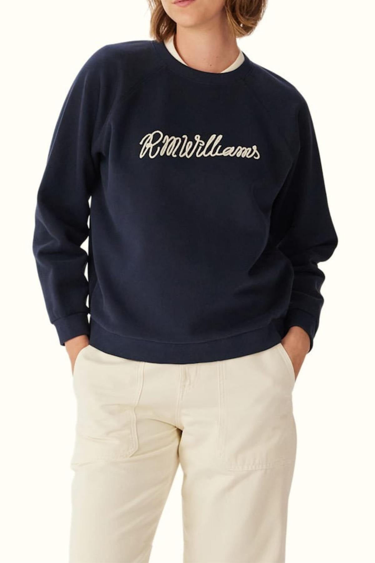 A person wearing the RM Williams Lasso Brushed Sweatshirt in navy with "R.M.Williams" embroidered in white script on the front and light-colored pants, standing with hands in pockets.