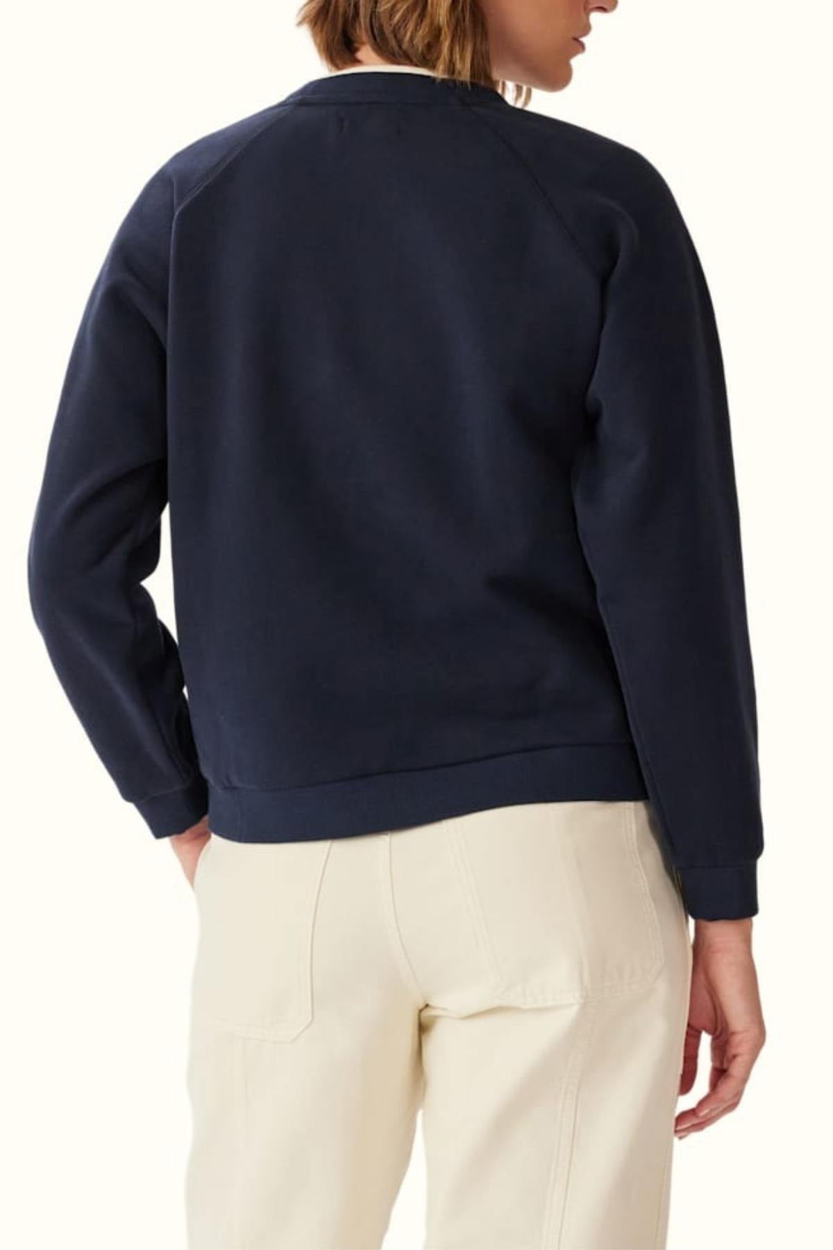 A person stands with their back to the camera, wearing a navy RM Williams Lasso Brushed Sweatshirt with branded embroidery and light pants. Their left hand rests on their hip.