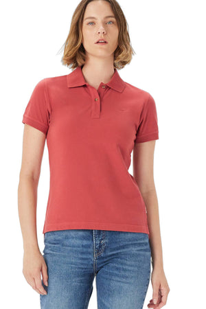 A woman with short light brown hair wears the RM Williams Margaret Polo, a red organic cotton shirt with longhorn embroidery, and blue jeans, standing against a white background.