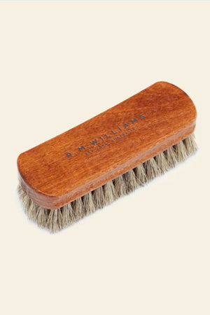 The RM Williams Medium Brush features a wooden handle engraved with "R.M.Williams" and light brown horse hair bristles, shown resting on a plain, light background.