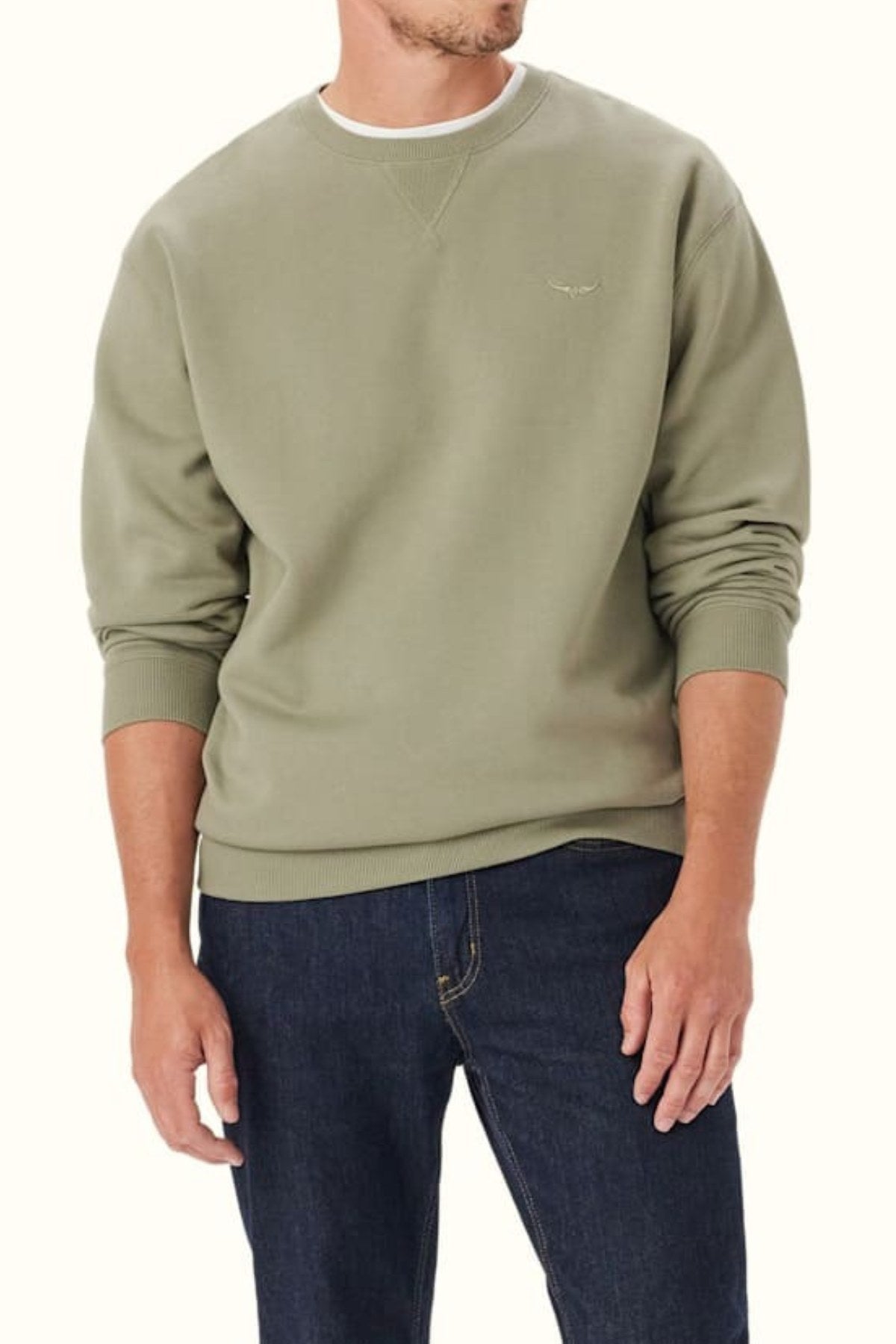 A man stands with his arms relaxed by his sides against a neutral background, wearing an RM Williams Mini Longhorn Sweatshirt in light olive green organic cotton and dark blue jeans.