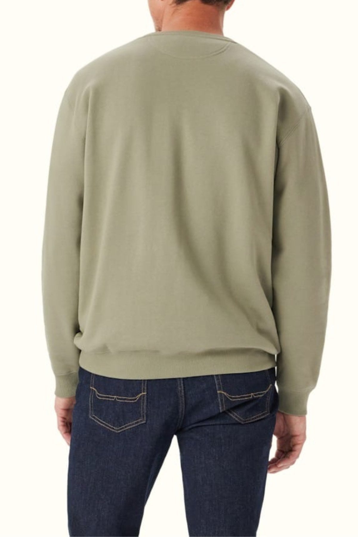 A person is shown from the back wearing the RM Williams Mini Longhorn Sweatshirt in light olive green with a ribbed crew neck, paired with dark blue jeans. The backdrop is plain and light-colored.