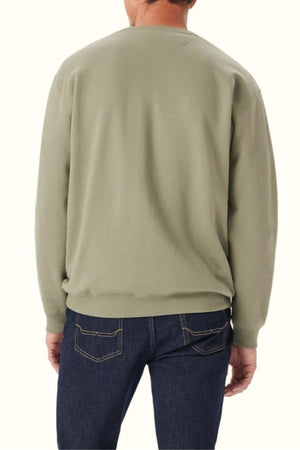 A person is shown from the back wearing the RM Williams Mini Longhorn Sweatshirt in light olive green with a ribbed crew neck, paired with dark blue jeans. The backdrop is plain and light-colored.