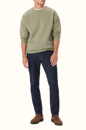 A man wears the RM Williams Mini Longhorn Sweatshirt in light olive green organic cotton with a ribbed crew neck, paired with dark blue jeans and brown suede shoes, standing against a plain light background.