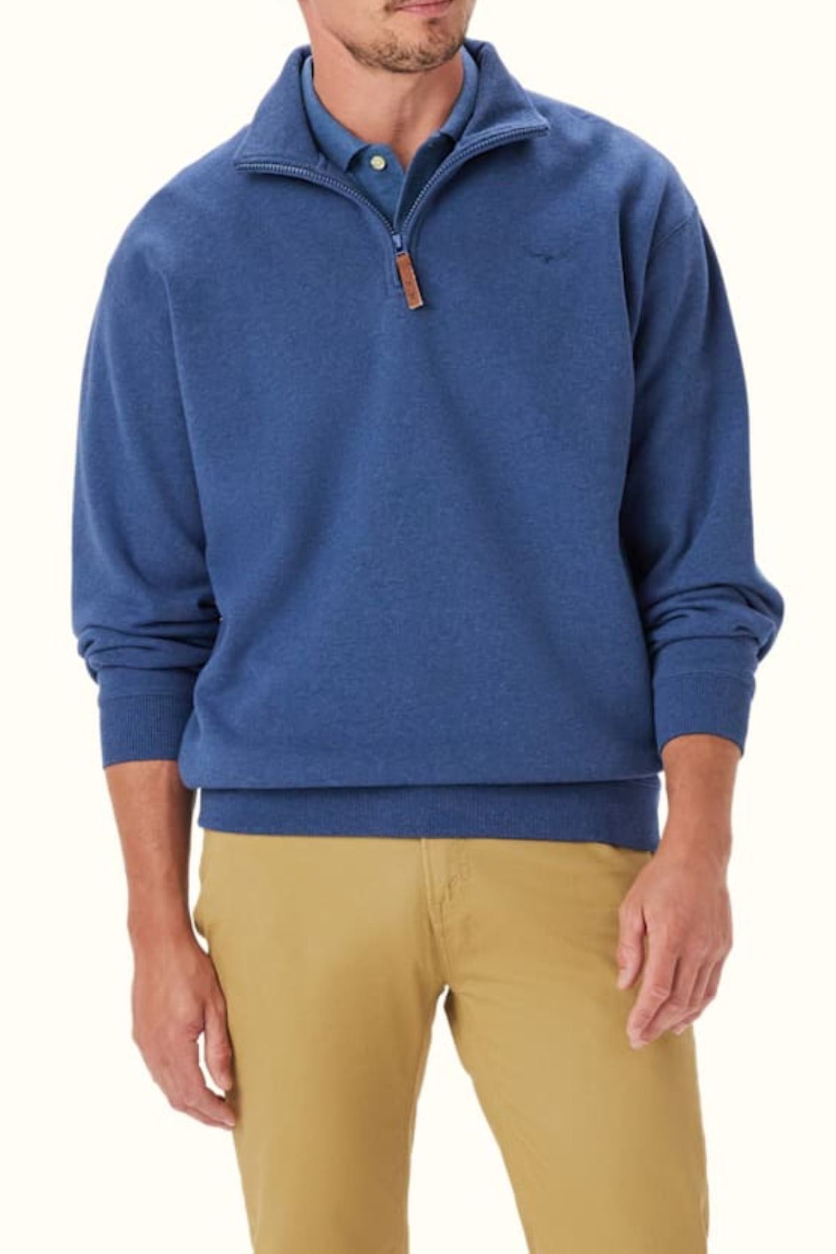 A man wears the RM Williams Mulyungarie Fleece, an organic cotton blue quarter zip pullover with Longhorn embroidery and a leather zipper pull, over a light blue collared shirt and tan pants, standing against a plain light background.