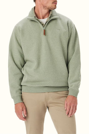A man in the RM Williams Mulyungarie Fleece—a light green organic cotton blend quarter-zip with a leather zipper pull and subtle Longhorn embroidery—pairs it with khaki pants, standing against a plain background, head turned slightly to the side.