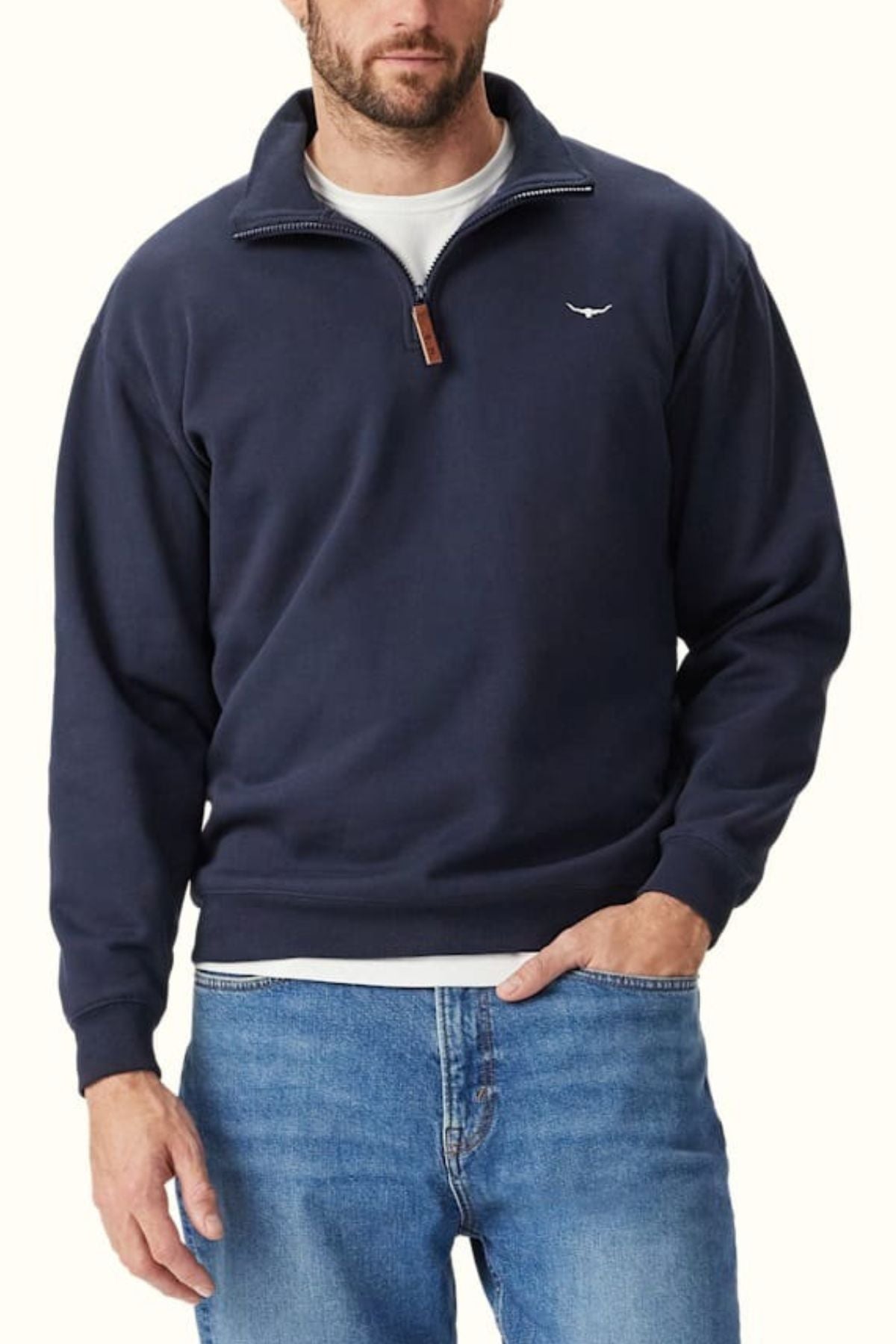 A man wears an RM Williams Mulyungarie Fleece, a navy blue organic cotton quarter-zip pullover with a small white chest logo, over a white undershirt and blue jeans. He stands against a light background with one hand in his pocket.