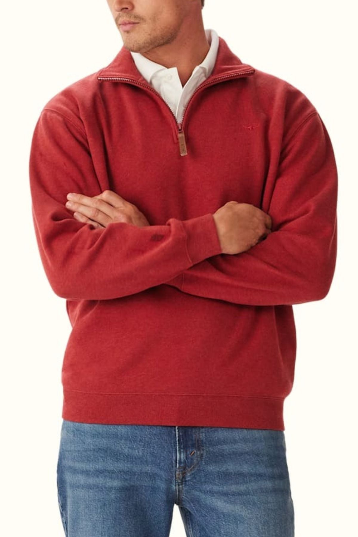 A man stands with arms crossed against a plain background, wearing the RM Williams Mulyungarie Fleece—a red organic cotton quarter zip pullover over a white collared shirt and blue jeans.
