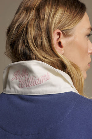 A woman with wavy blonde hair is seen from behind, wearing the RM Williams Narran Heritage 3 Panel Rugby in blue with a white collar turned up, showing pink heritage logo embroidery reading "R.M. Williams.