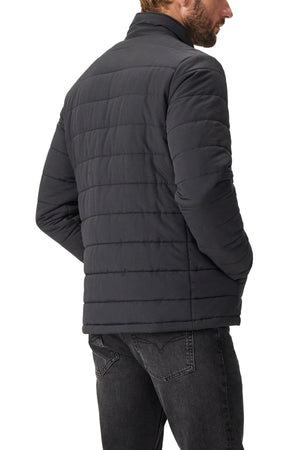 A man stands with his back to the camera, hands in pockets, wearing the RM Williams Patterson Creek Jacket—a black lightweight quilted coat made from recycled nylon—and dark jeans, set against a plain white background.