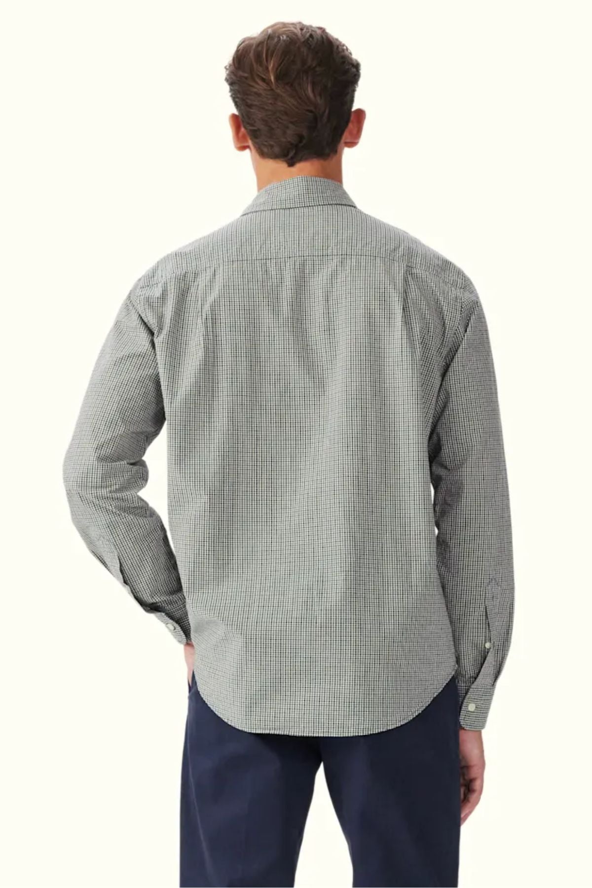 A man is seen from behind wearing the RM Williams Quinn Classic Poplin Shirt, a long-sleeve, yarn-dyed check shirt in black and white, paired with navy pants. His right hand rests on his hip against a plain off-white background.