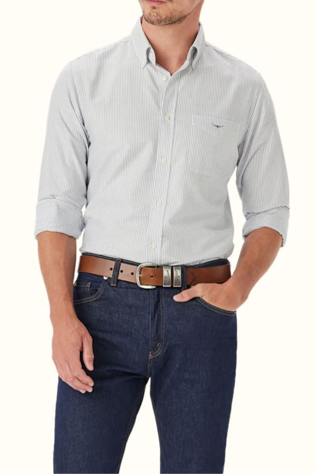 A man wears the RM Williams Regular Oxford Stripe Shirt in light blue and white stripes, sleeves rolled up, paired with dark blue jeans and a brown belt. The shirt has a chest pocket, and his head is partially out of frame.