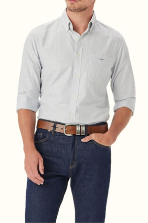 A man wears the RM Williams Regular Oxford Stripe Shirt in light blue and white stripes, sleeves rolled up, paired with dark blue jeans and a brown belt. The shirt has a chest pocket, and his head is partially out of frame.