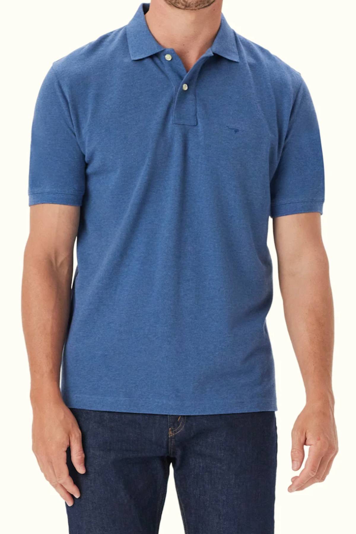 A man wears the RM Williams Rod Polo, a short-sleeve, blue organic cotton shirt with a collar, two top buttons, and subtle Longhorn embroidery, paired with dark blue jeans against a plain light background.