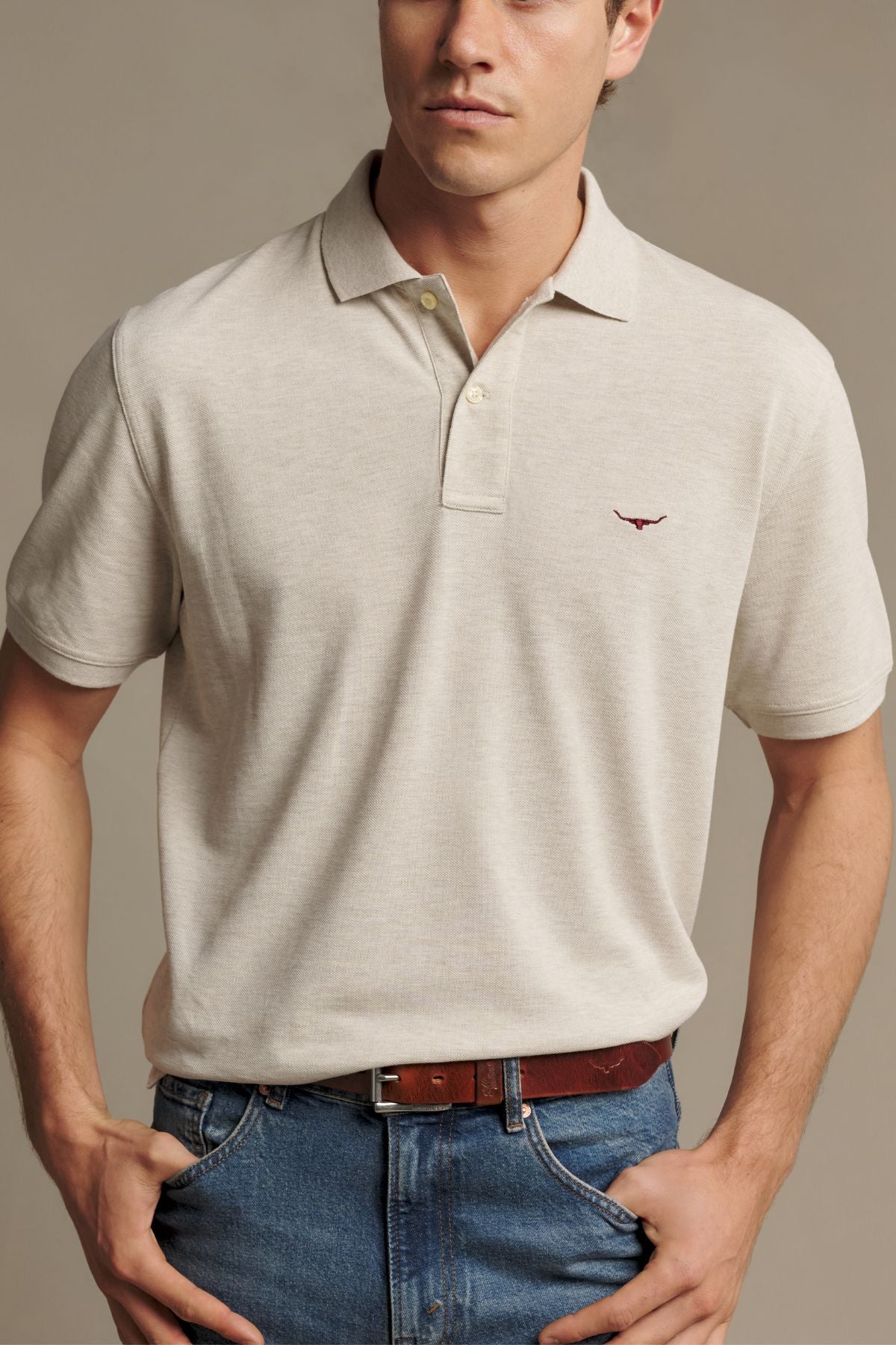 A man wearing the RM Williams Rod Polo in light beige Australian cotton, featuring a small red Longhorn embroidery, paired with blue jeans and a brown belt, stands with his hands in his pockets against a neutral background.