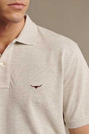 A man is seen wearing the RM Williams Rod Polo in light beige, made from premium Australian cotton and featuring a small maroon longhorn embroidery on the left chest. Only the lower half of his face and upper torso are visible.