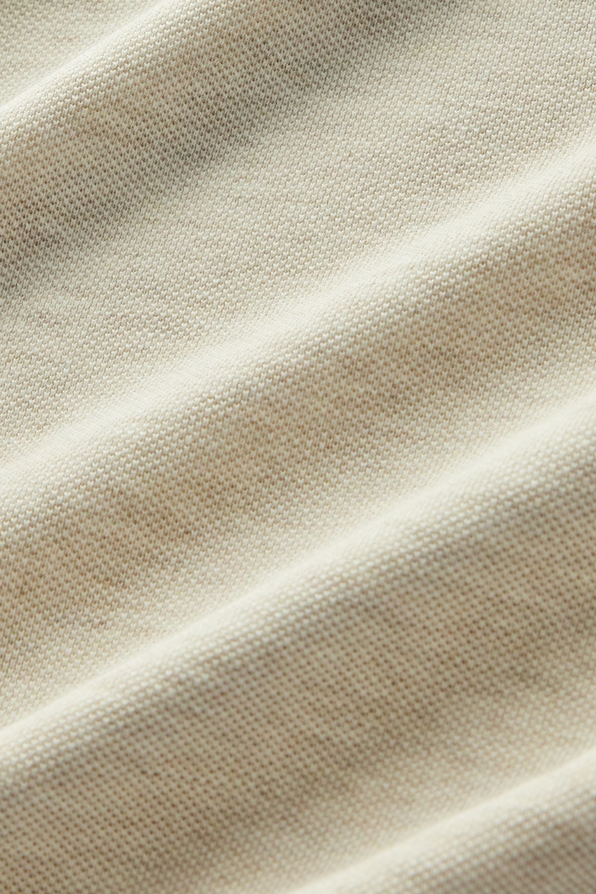 Close-up of beige fabric showcasing a visible knit texture with gentle, diagonal folds that create subtle shadows and highlights—evoking the premium materials found in the RM Williams Rod Polo.