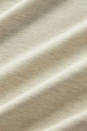 Close-up of beige fabric showcasing a visible knit texture with gentle, diagonal folds that create subtle shadows and highlights—evoking the premium materials found in the RM Williams Rod Polo.