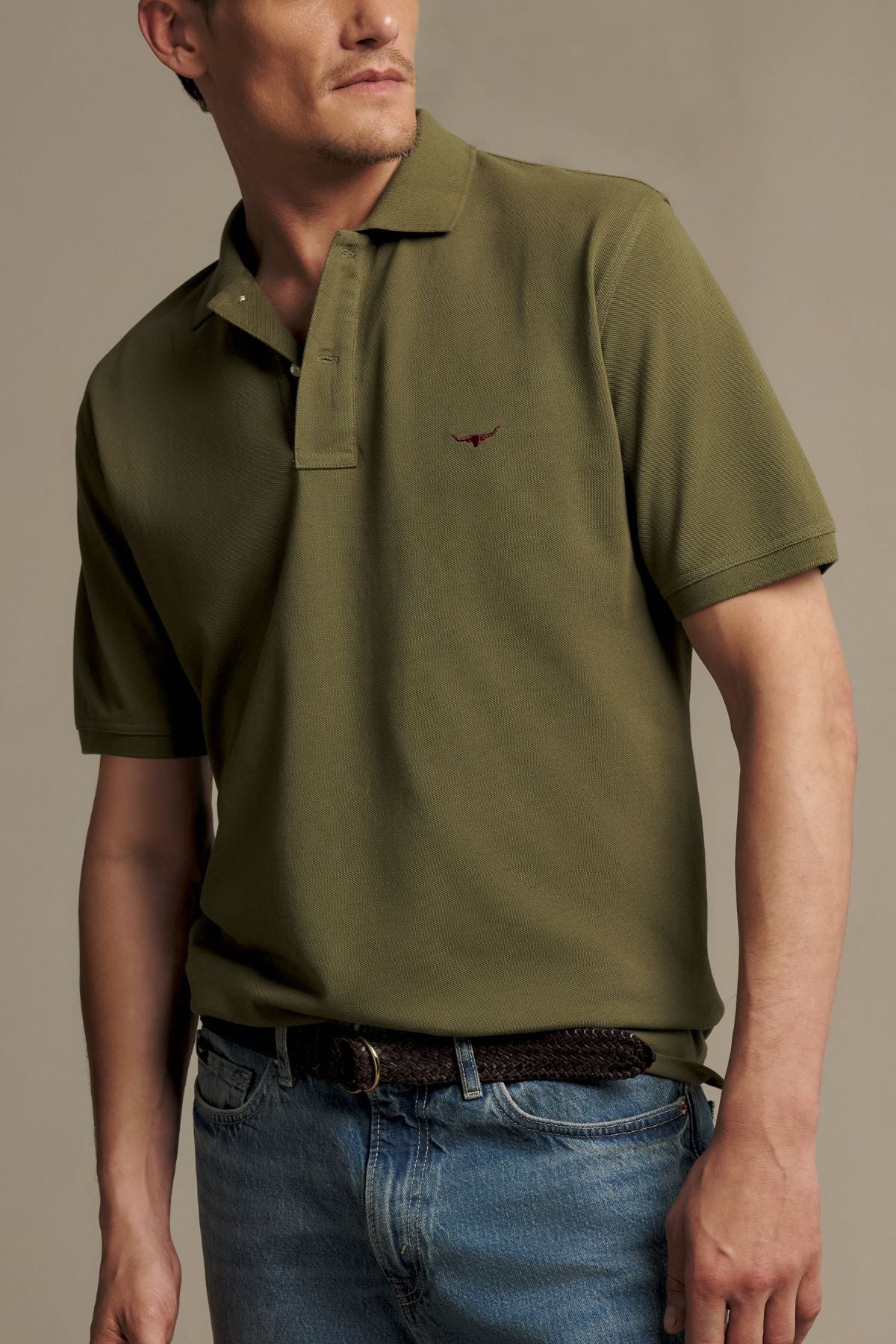 A man wearing the RM Williams Rod Polo in olive green—an Australian cotton short-sleeve shirt with a ribbed collar—paired with blue jeans and a woven brown belt, facing slightly to the left against a neutral background.