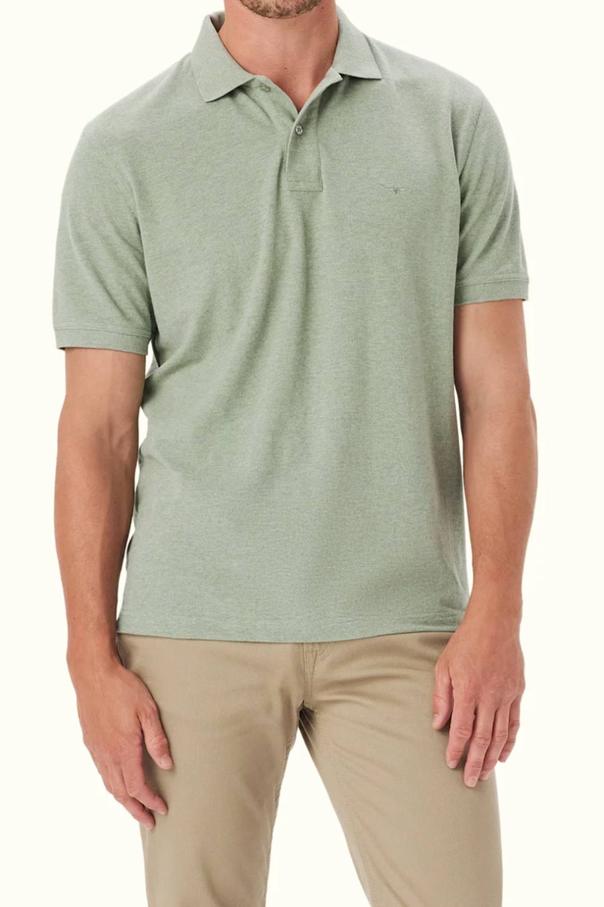 A man wearing the RM Williams Rod Polo in light green organic cotton, featuring subtle Longhorn embroidery, with beige pants stands against a plain light background. His face is partially obscured.