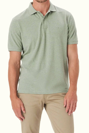 A man wearing the RM Williams Rod Polo in light green organic cotton, featuring subtle Longhorn embroidery, with beige pants stands against a plain light background. His face is partially obscured.