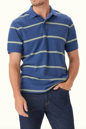 A man in the RM Williams Rod Polo—short-sleeved, blue with light green stripes, organic cotton—and dark blue jeans stands with his left hand in his pocket against a plain background.