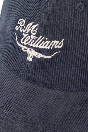 A close-up of the RM Williams Script Longhorn Cord Cap, a dark organic cotton corduroy hat featuring white "R.M. Williams" embroidery above a stylized longhorn logo.