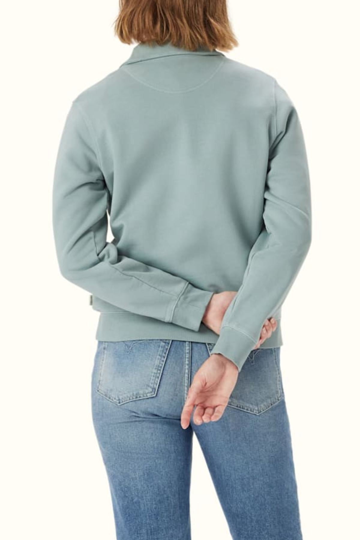 A person with light brown hair, wearing the RM Williams Trickett Zip Neck Sweatshirt in light blue with Longhorn embroidery and blue jeans, stands facing away from the camera, their left hand tucked behind their back.