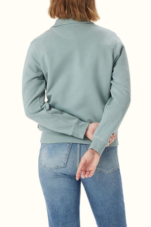 A person with light brown hair, wearing the RM Williams Trickett Zip Neck Sweatshirt in light blue with Longhorn embroidery and blue jeans, stands facing away from the camera, their left hand tucked behind their back.