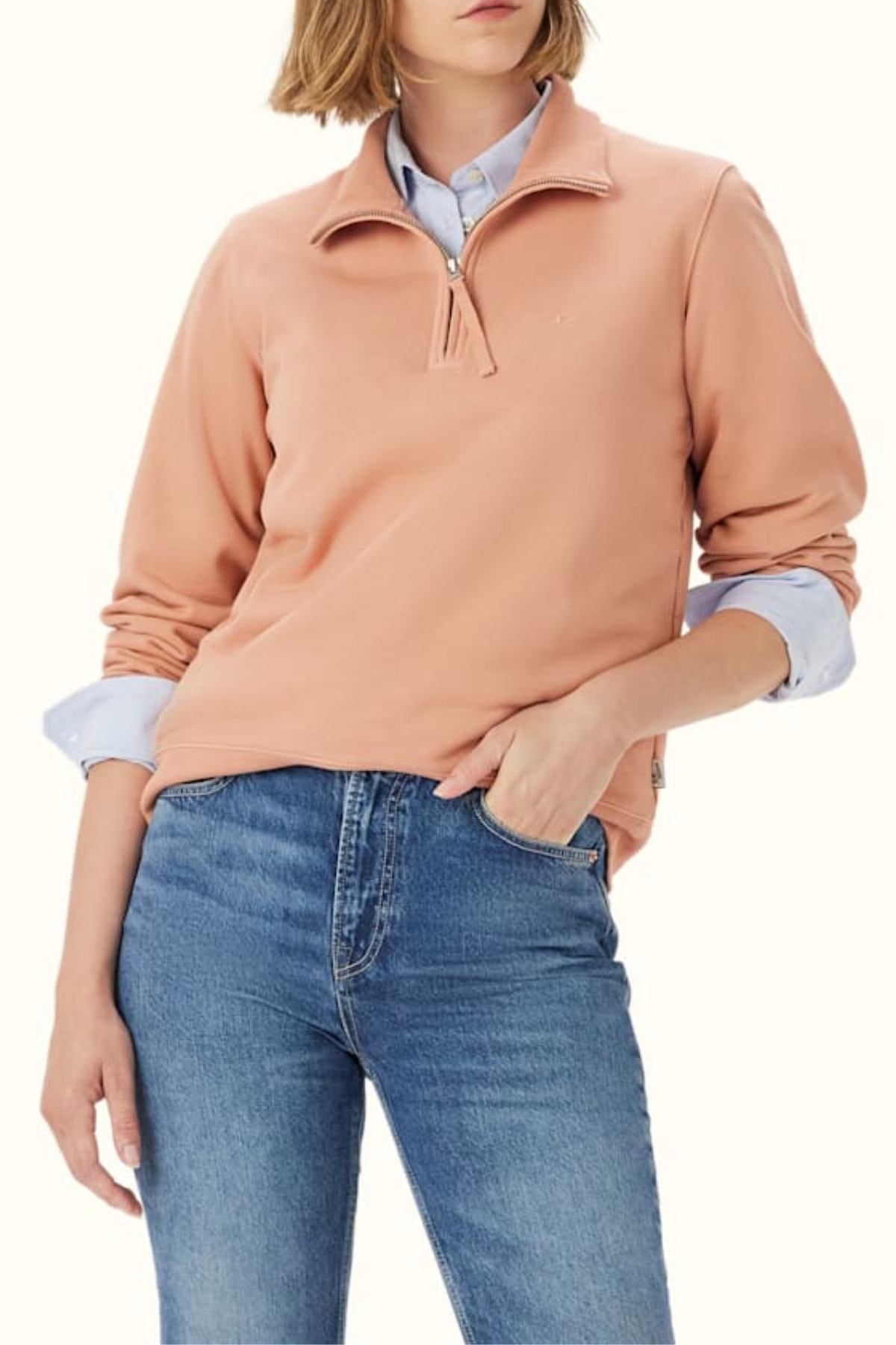 A woman wears the RM Williams Trickett Zip Neck Sweatshirt in light peach over a light blue collared shirt and blue jeans, standing with one hand in her pocket against a plain background.