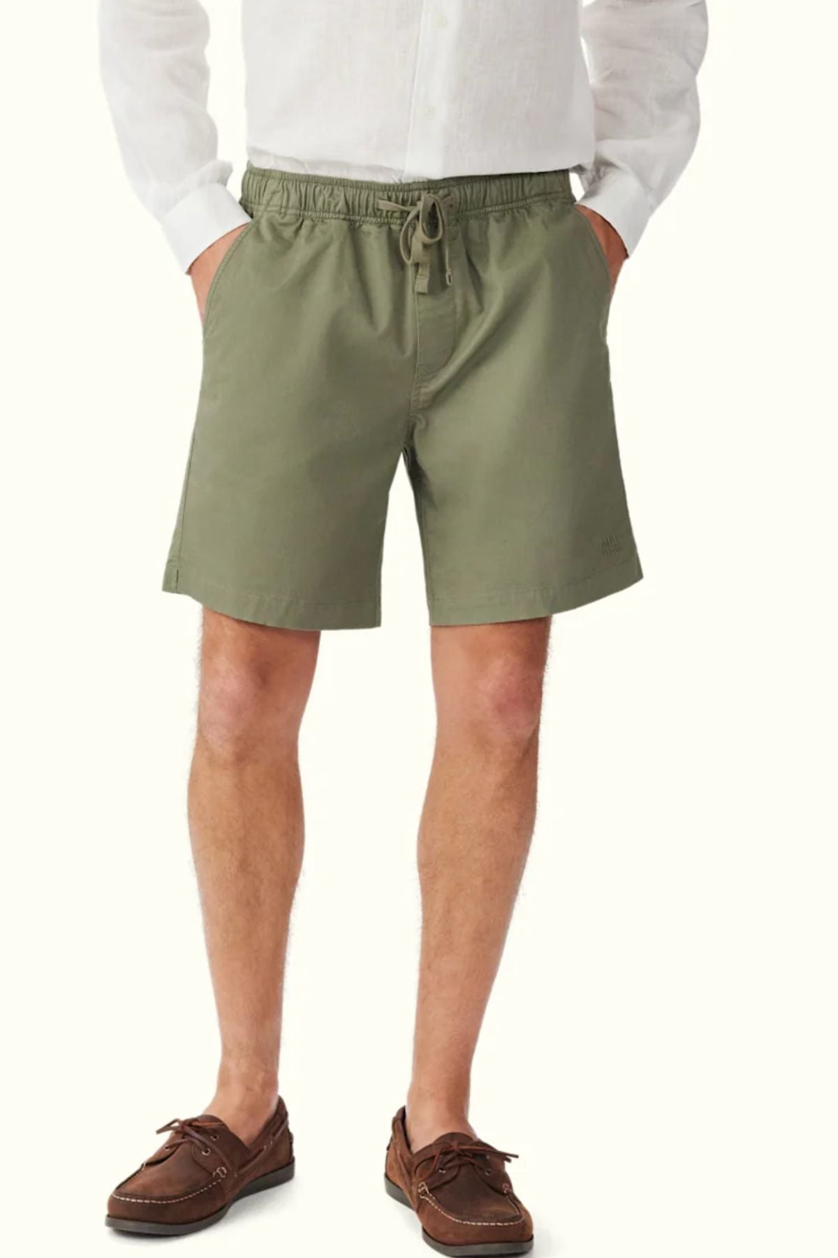 A person stands with hands in pockets against a light background, wearing RM Williams Washed Rugby Shorts, a white long-sleeve shirt, and brown boat shoes.