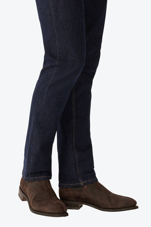 A person stands against a plain light background, wearing dark blue jeans and RM Williams Comfort Craftsman Boots in brown waxed suede with Goodyear welt construction. Only the lower legs and feet are visible.