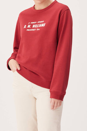 Someone wearing the RM Williams Heritage Brushed Sweatshirt in red with white embroidered text and off-white pants stands with one hand in their pocket. The sweatshirt features “5 Percy Street R.M. Williams Prospect S.A.”.