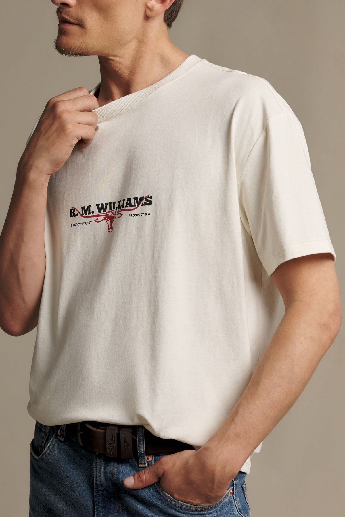 A man wearing the RM Williams Holt Tee—a cream T-shirt featuring “R. M. WILLIAMS” and a logo on the front—paired with blue jeans and a brown belt, stands against a neutral background.