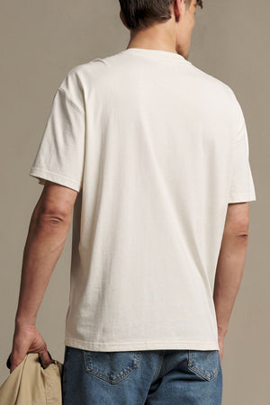 A man is shown from the back wearing the RM Williams Holt Tee in plain white and blue jeans, holding a beige jacket in his left hand against a neutral background.
