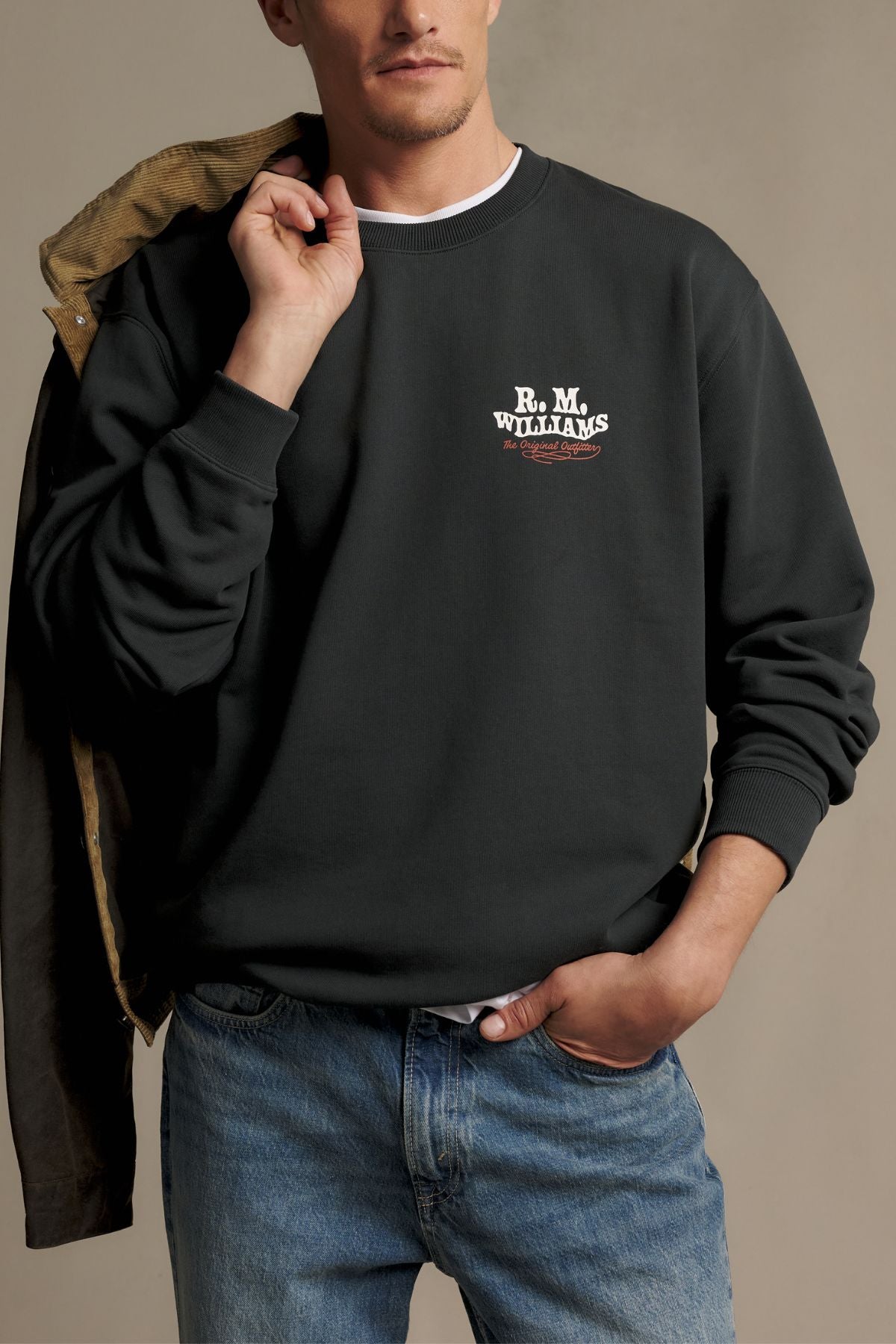 A man wearing an RM Williams Kandos Crew sweatshirt in black with blue jeans stands against a neutral background, holding a jacket over his shoulder with one hand tucked in his pocket.