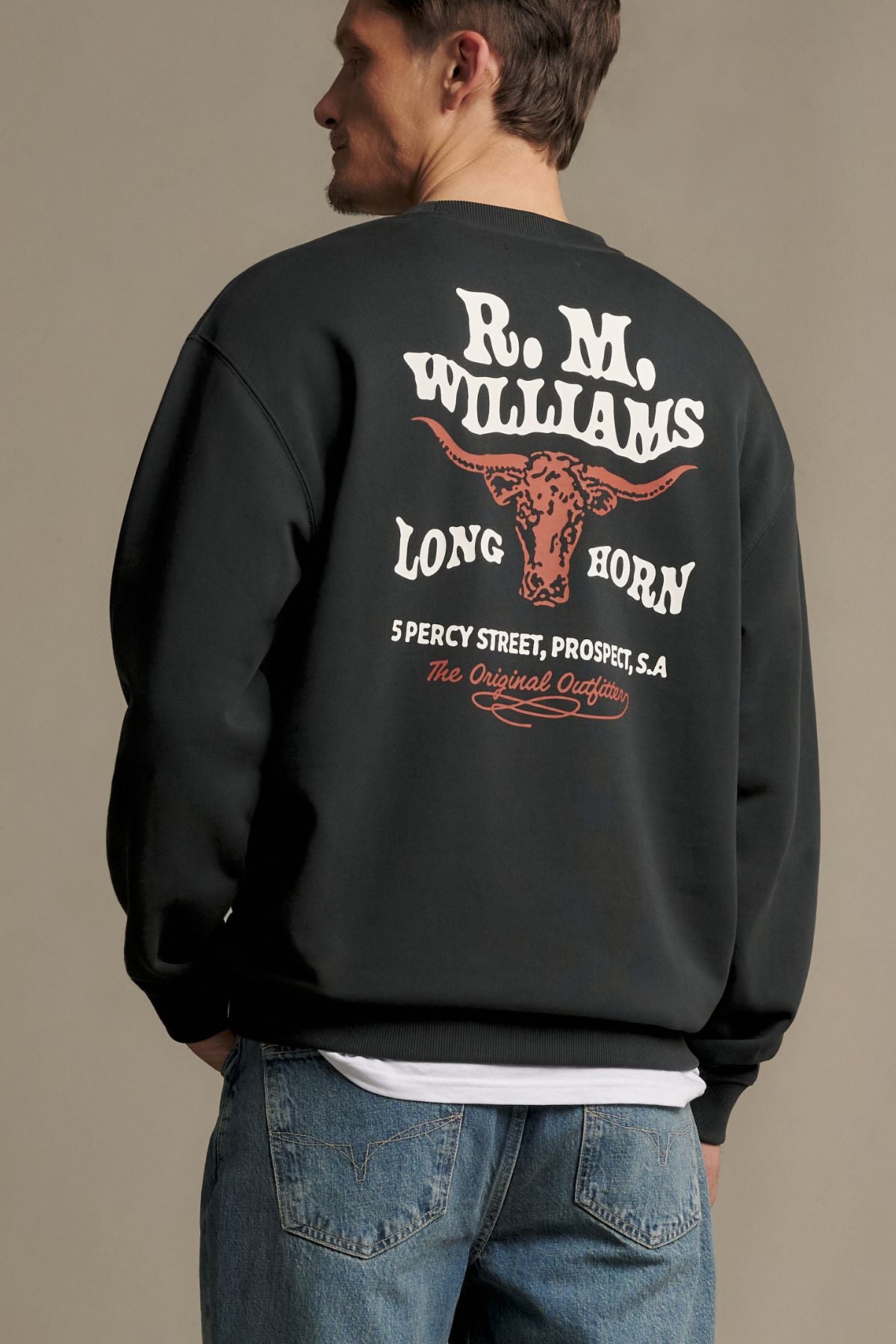 A man in blue jeans and an RM Williams Kandos Crew sweatshirt featuring “R.M. Williams Long Horn” and a bull graphic on the back stands facing away from the camera.