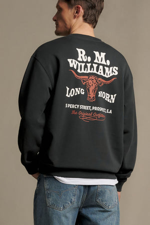 A man in blue jeans and an RM Williams Kandos Crew sweatshirt featuring “R.M. Williams Long Horn” and a bull graphic on the back stands facing away from the camera.