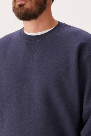 A bearded man wears the RM Williams Mini Longhorn Sweatshirt, a dark blue ribbed crew neck made from an organic cotton blend with subtle Longhorn embroidery on the chest over a white T-shirt. Only his lower face and upper torso are visible.