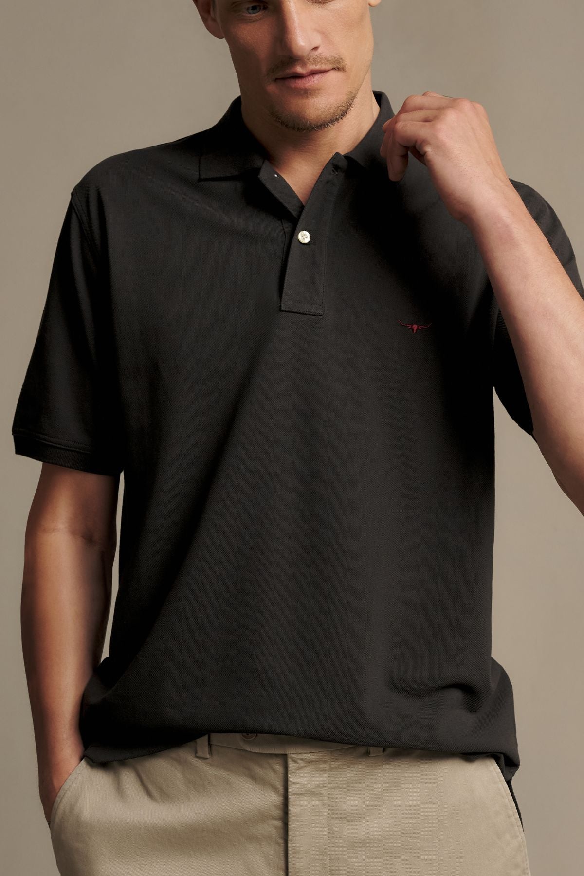 A man wearing the RM Williams Rod Polo in black paired with beige pants stands against a plain background, holding his shirt collar with one hand and looking slightly down.