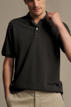 A man wearing the RM Williams Rod Polo in black paired with beige pants stands against a plain background, holding his shirt collar with one hand and looking slightly down.
